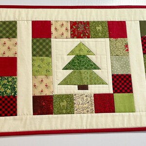 May include: A quilted Christmas table runner featuring three central squares with green Christmas tree designs. The trees are surrounded by red, green, and cream-colored patchwork squares. The runner has a cream-colored border with a red trim.