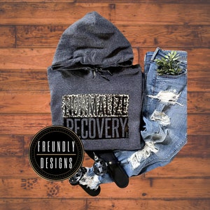 May include: A gray hooded sweatshirt with the text "NORMALIZE RECOVERY" printed on the front. The text is in black with a leopard print background. The sweatshirt is laying on a wooden surface with a pair of jeans and a black shoe.