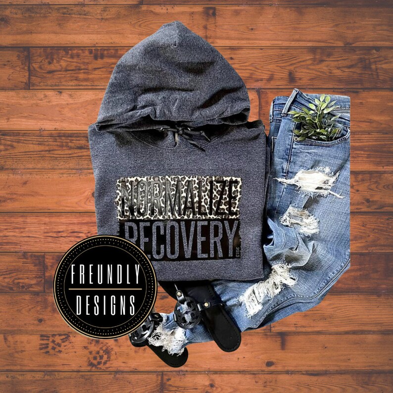May include: A gray hooded sweatshirt with the text "NORMALIZE RECOVERY" printed on the front. The text is in black with a leopard print background. The sweatshirt is laying on a wooden surface with a pair of jeans and a black shoe.