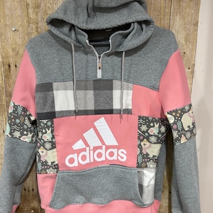 Könnte beinhalten: Grau-rosa Adidas Kapuzenpullover mit Fronttasche und Kapuze. Der Hoodie hat ein Karomuster und Aufnäher mit Hasen und Bären. Das Adidas-Logo ist prominent in Weiß dargestellt.