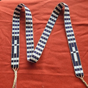 May include: A white and blue beaded strap with a checkered pattern. The strap is made of small beads and has a tassel at each end.