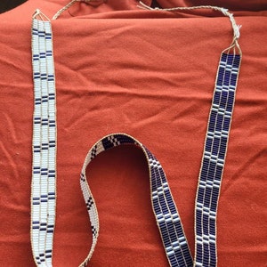May include: A white and dark blue beaded strap with a geometric pattern. The strap is made of small, round beads and is approximately 1.5 inches wide.