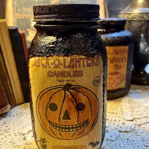 May include: A vintage-style Halloween candle jar with a black lid and a label that reads "Jack-O-Lantern Candles." The label features a smiling pumpkin face. The jar is part of a set of 10 and has a price tag of 29 cents.