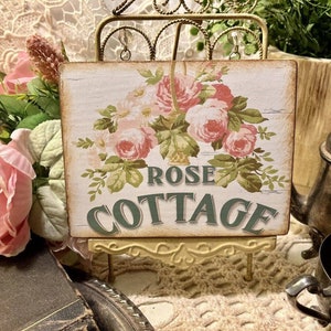 May include: A white wooden sign with a floral design and the text "ROSE COTTAGE" in green letters. The sign is on a small gold metal easel.