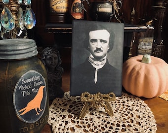 May include: A black and white portrait of Edgar Allan Poe on a black background, framed and displayed on a gold metal stand. The stand is on a white crocheted doily. The portrait is surrounded by various Halloween-themed decorations, including a mason jar with a black and orange sticker that reads "Something Wicked Comes This Way", a pumpkin, and a bottle of liquid with a label that reads "Witch's Brew".