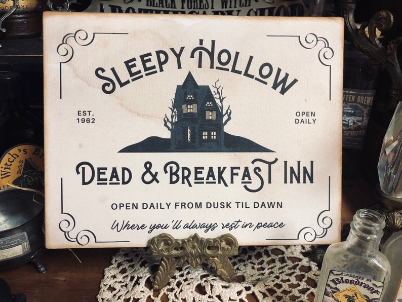 May include: A white wooden sign with black lettering that reads "Sleepy Hollow Dead & Breakfast Inn" with a black silhouette of a house with windows. The sign also says "Est. 1962" and "Open Daily From Dusk Til Dawn Where you'll always rest in peace".