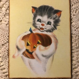 Pu&ograve; includere: Un'illustrazione vintage di Halloween di un gatto grigio e bianco che tiene una zucca arancione intagliata. Il gatto indossa un costume da fantasma bianco e ha un'espressione felice sul viso.