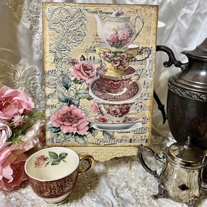 May include: A vintage-inspired teacup and saucer set with floral designs. The set is stacked on top of a music sheet with the words "Favorite Germany" printed on it. The teacups are decorated with pink and red roses and green leaves. The set is displayed on a wooden stand.