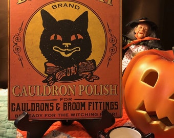 May include: A vintage-style sign with a black cat illustration and the text "BLACK CAT BRAND CAULDRON POLISH FOR CAULDRONS & BROOM FITTINGS BE READY FOR THE WITCHING HOUR!" The sign is on a black easel and a black cat candle holder is in the foreground.