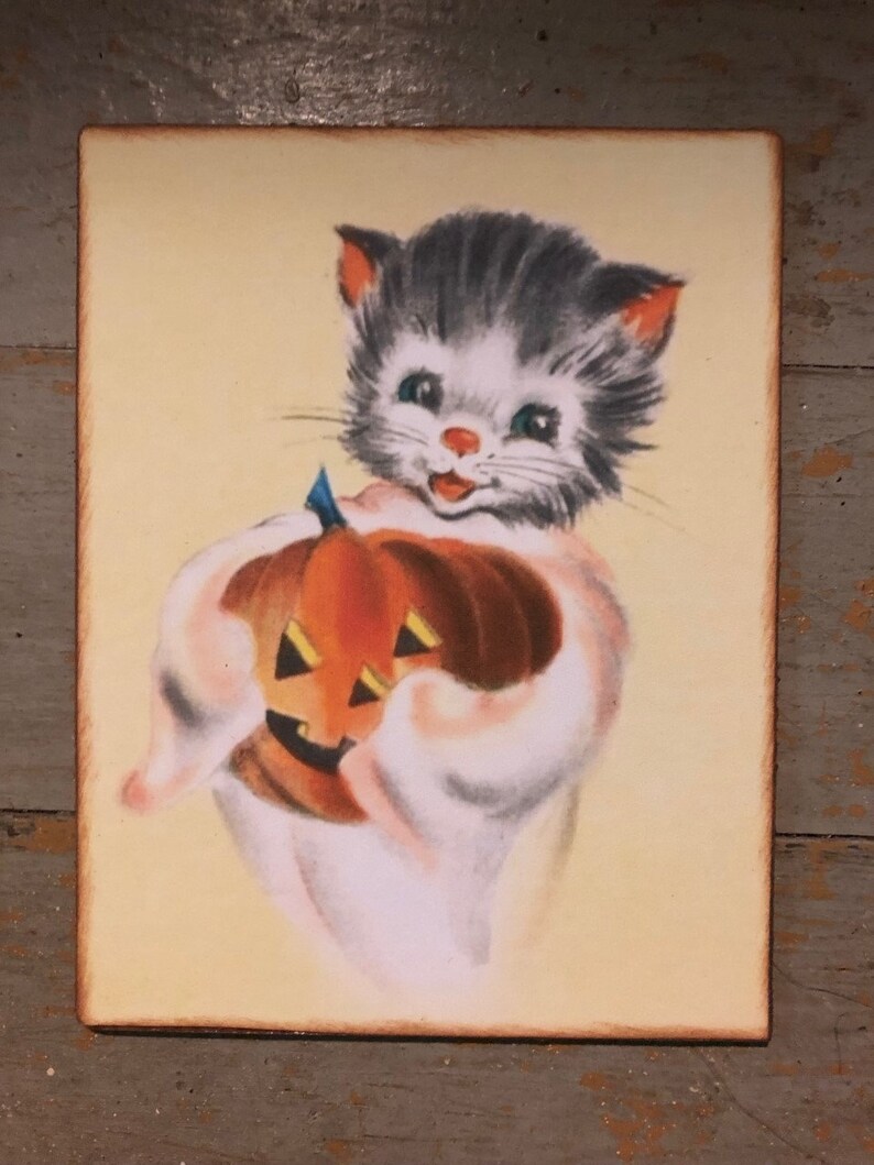 Pu&ograve; includere: Un'illustrazione vintage di Halloween di un gattino grigio e bianco che tiene una zucca arancione intagliata. Il gattino indossa un costume da fantasma bianco e ha un'espressione felice sul viso.