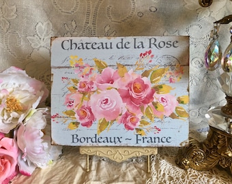 May include: A white wooden sign with a distressed finish featuring a watercolor painting of pink roses and the text "Chateau de la Rose Bordeaux - France".