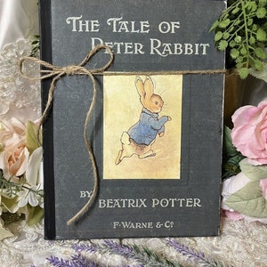 May include: A vintage copy of "The Tale of Peter Rabbit" by Beatrix Potter. The book has a dark gray cover with white lettering and a color illustration of Peter Rabbit. A twine bow wraps around the book, which is surrounded by flowers and greenery.