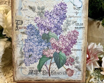 May include: A vintage-style sign with a floral design featuring purple and pink lilac blossoms. The sign reads "Fleur de Lilas" and "Paris, 1889" with the text "St. Paul Charvet, Rue des Fabriques" at the bottom.