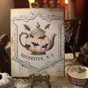 May include: A vintage-style sign featuring a teapot design with bats, set against a pink and white striped background. The sign includes the text "Rochester, N.Y." and is displayed on a small brass stand, evoking a Halloween or gothic theme.