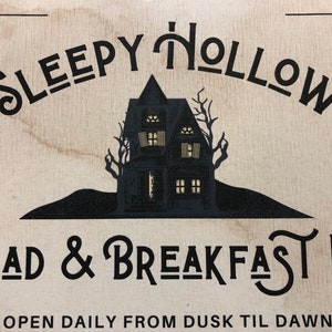 May include: A vintage-style sign for a bed and breakfast called "Sleepy Hollow Dead & Breakfast Inn". The sign features a black silhouette of a spooky house with lit windows against a beige background. The text "EST. 1962" and "OPEN DAILY FROM DUSK TIL DAWN" is also included.