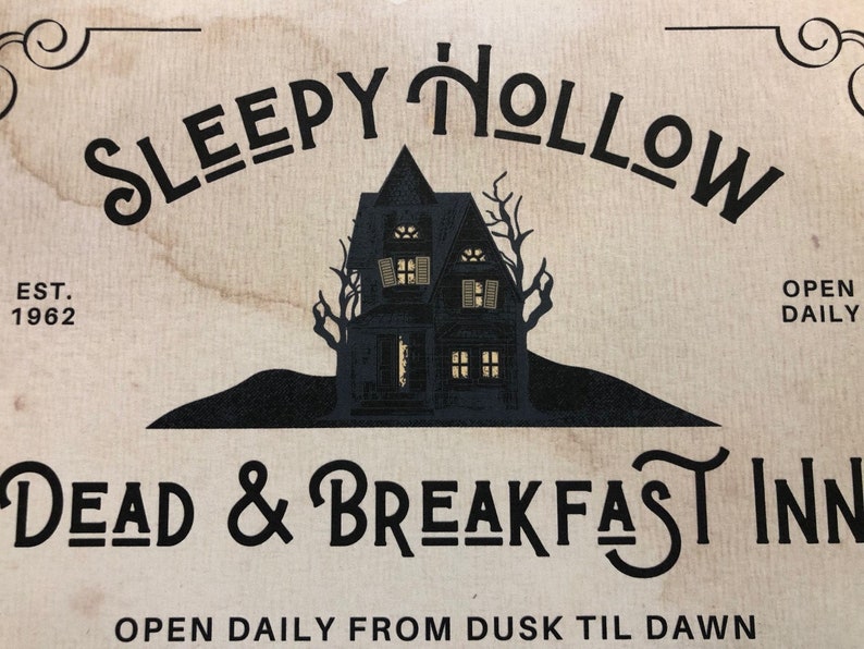May include: A vintage-style sign for a bed and breakfast called "Sleepy Hollow Dead & Breakfast Inn". The sign features a black silhouette of a spooky house with lit windows against a beige background. The text "EST. 1962" and "OPEN DAILY FROM DUSK TIL DAWN" is also included.