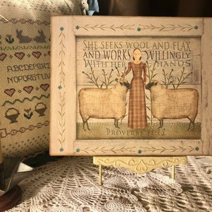 May include: A framed print with a white background and a woman in a red and white plaid dress standing between two sheep. The text reads "She seeks wool and flax and works willingly with her hands" and "Proverbs 31:13".