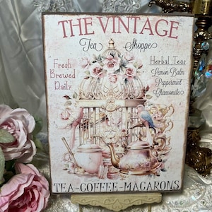 May include: A vintage-style sign with the text "THE VINTAGE Tea Shoppes" in pink lettering. The sign features a birdcage with pink roses and two birds, one blue and one pink. The sign also lists "Fresh Brewed Daily" and "Herbal Teas: Lemon Balm, Peppermint, Chamomile". The bottom of the sign reads "TEA-COFFEE-MACARONS".