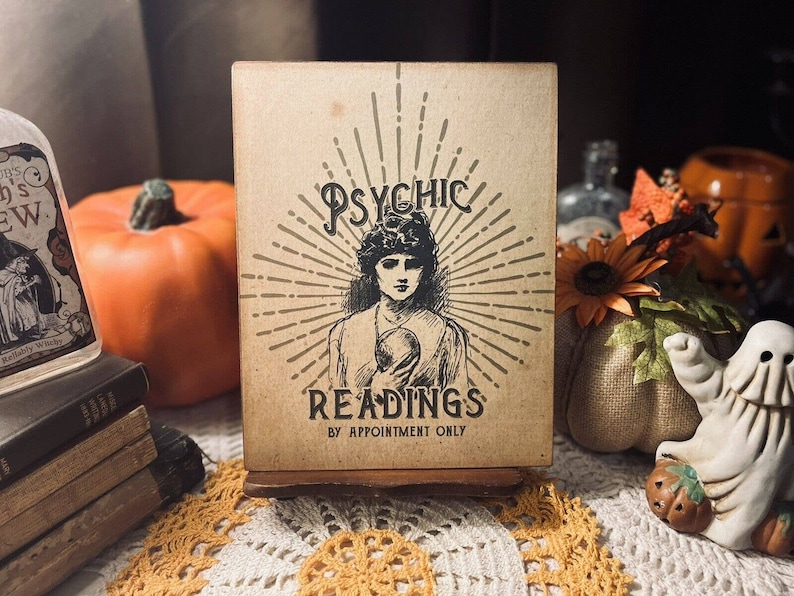 May include: A vintage-style sign with a brown background and gold accents reads "Psychic Readings by Appointment Only". The sign features a black and white illustration of a woman with long hair and a flowing dress. The sign is sitting on a table with a white tablecloth and fall decorations.