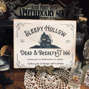 May include: A white sign with black lettering that reads "Sleepy Hollow Est. 1962 Dead & Breakfast Inn Open Daily From Dusk Til Dawn Where you'll always rest in peace". The sign features a black silhouette of a house with a single window and a tree in front of it.