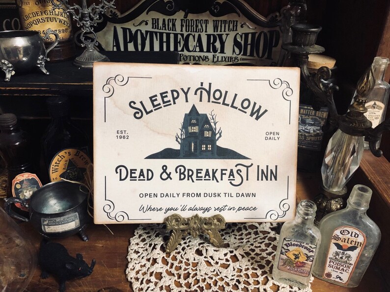 May include: A white sign with black lettering that reads "Sleepy Hollow Est. 1962 Dead & Breakfast Inn Open Daily From Dusk Til Dawn Where you'll always rest in peace". The sign features a black silhouette of a house with a single window and a tree in front of it.