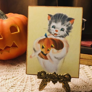 Pu&ograve; includere: Un'illustrazione vintage a tema Halloween con un gatto bianco che tiene una zucca intagliata. Il gatto ha il pelo nero e grigio e gli occhi azzurri. L'illustrazione &egrave; su uno sfondo giallo ed &egrave; incorniciata in una cornice di legno.
