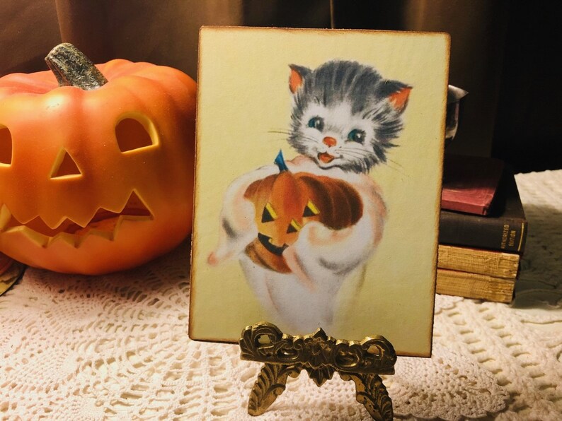 Pu&ograve; includere: Un'illustrazione vintage a tema Halloween con un gatto bianco che tiene una zucca intagliata. Il gatto ha il pelo nero e grigio e gli occhi azzurri. L'illustrazione &egrave; su uno sfondo giallo ed &egrave; incorniciata in una cornice di legno.