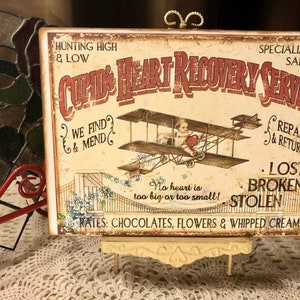 May include: Vintage-style sign with the text "Cupid's Heart Recovery Service" and an illustration of a biplane. The sign has a distressed look with text like "Hunting High & Low" and "Repair & Return". It is displayed on a small, decorative easel.