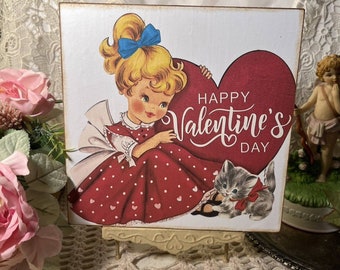 Può includere: Un biglietto di San Valentino vintage con una ragazza in un vestito rosso a pois bianchi che tiene un grande cuore rosso su cui è scritto "Happy Valentine's Day". Un gattino grigio con un fiocco rosso è seduto accanto a lei.