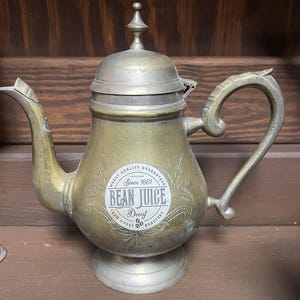 May include: Antique brass coffee pot with a curved handle and spout. The pot features the text "BEAN JUICE Decaf Roastery" and "Worst Quality Guaranteed Since 1661". The pot has a decorative finial on the lid.