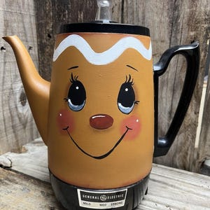 May include: A brown General Electric coffee pot with a gingerbread man face painted on it. The pot has a black base and handle, with the text "MILD MED STRONG" on the front. The pot has a clear lid.