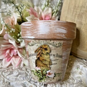 May include: A whitewashed terracotta flower pot with a vintage-style design featuring a brown rabbit and pink flowers. The pot has the text "Antiquites Aux De Bords" printed on it.
