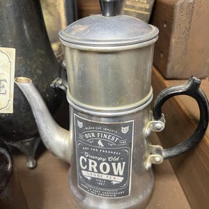 May include: A vintage silver-toned tea pot with a black handle and lid. The pot features a label that reads "Grumpy Old Crow Loose Tea" and "Black Cat Imports." The design has a classic, antique aesthetic, perfect for tea enthusiasts.