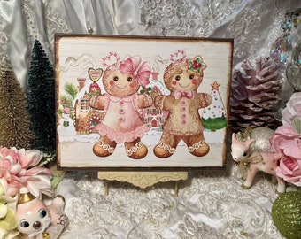 May include: A wooden sign with a painted image of a gingerbread couple. The woman is wearing a pink dress and the man is wearing a brown suit. They are standing in front of a gingerbread house with a snowy background.