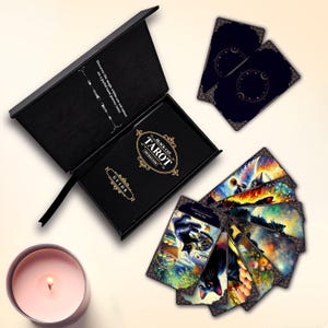 May include: A black box with a gold inscription that reads "Black Cat Tarot" and "Guide" on the front. The box is open, revealing a deck of tarot cards with black cats and colorful illustrations. The cards are fanned out on a white surface.