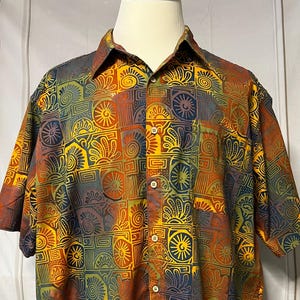 May include: Short-sleeved button-down shirt with a vibrant, geometric pattern in shades of orange, yellow, blue, and green. The shirt is displayed on a mannequin, showcasing its colorful design and casual style.
