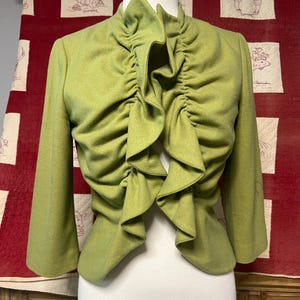 May include: A light green jacket with a ruffled front and three-quarter sleeves. The jacket is displayed on a mannequin against a red background with embroidered squares. The jacket's design features a high, ruffled collar.