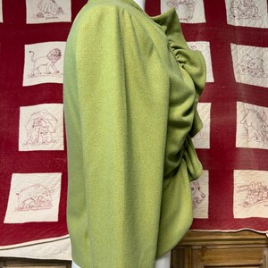 Może przedstawiać: Jasnozielona kurtka w stylu vintage z falbanowym kołnierzem i dopasowanym krojem. Kurtka jest prezentowana na manekinie, prezentując jej teksturowaną tkaninę i detale projektu. Tło przedstawia czerwoną kołdrę z haftowanymi kwadratami.