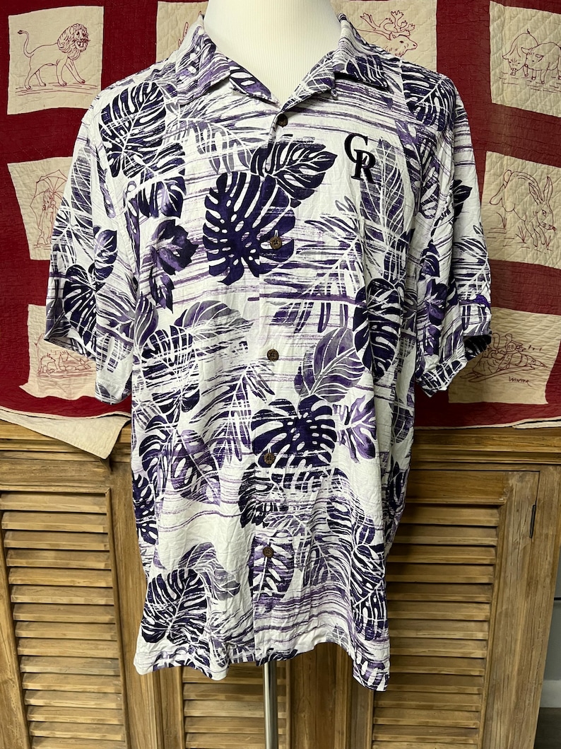 May include: White button-up shirt with a purple leaf pattern. The shirt has short sleeves and a classic collar. The shirt has a logo on the chest. The shirt is displayed on a mannequin.