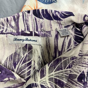 May include: Close-up of a Tommy Bahama button-down shirt with a purple and grey leaf pattern on a cream background. The shirt is labelled XL/TG and "Fabriqu&eacute; en Chine". Another shirt with a blue and orange floral pattern is partially visible.