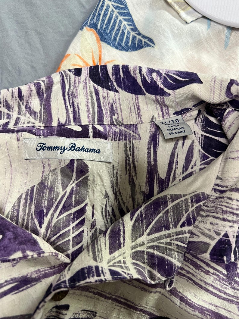 May include: Close-up of a Tommy Bahama button-down shirt with a purple and grey leaf pattern on a cream background. The shirt is labelled XL/TG and "Fabriqu&eacute; en Chine". Another shirt with a blue and orange floral pattern is partially visible.
