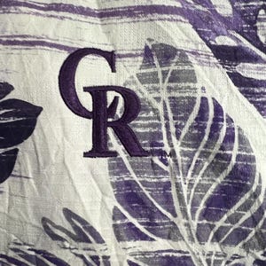 May include: Close-up of a white and purple patterned fabric. The fabric features a repeating leaf design and the embroidered letters "CR" in purple. The fabric has a textured appearance.