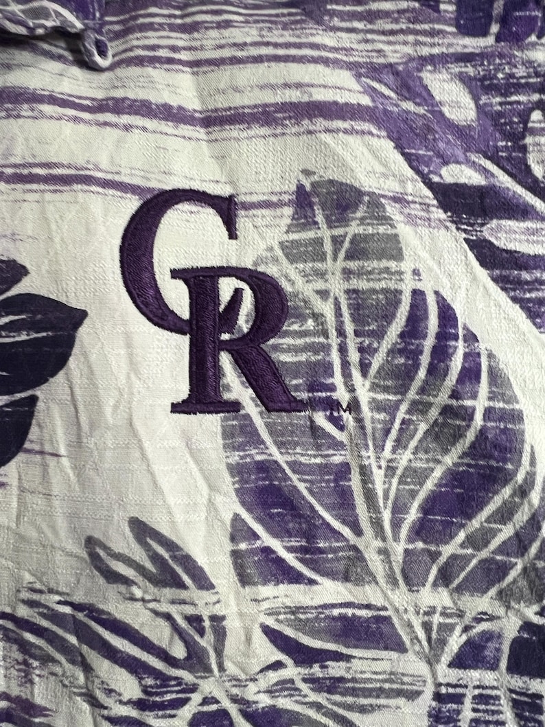 May include: Close-up of a white and purple patterned fabric. The fabric features a repeating leaf design and the embroidered letters "CR" in purple. The fabric has a textured appearance.