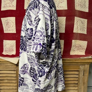 May include: A short-sleeved shirt with a white base, featuring a bold pattern of purple leaves. The shirt is displayed on a mannequin, highlighting its loose fit and tropical design. The fabric appears lightweight. The shirt's design is reminiscent of a Hawaiian shirt.