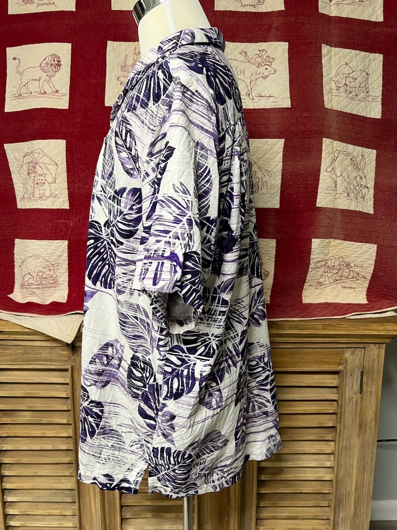 May include: A short-sleeved shirt with a white base, featuring a bold pattern of purple leaves. The shirt is displayed on a mannequin, highlighting its loose fit and tropical design. The fabric appears lightweight. The shirt's design is reminiscent of a Hawaiian shirt.