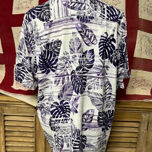 May include: White button-down shirt with a purple and navy leaf pattern. The short-sleeved shirt features a tropical design with large leaves and horizontal stripes. The shirt is displayed on a mannequin.