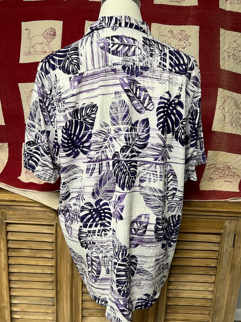 May include: White button-down shirt with a purple and navy leaf pattern. The short-sleeved shirt features a tropical design with large leaves and horizontal stripes. The shirt is displayed on a mannequin.