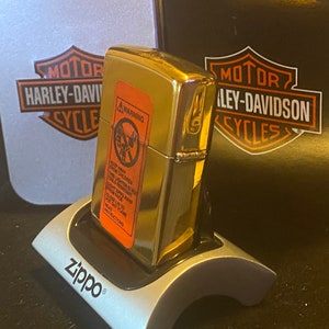 May include: A gold-tone Zippo lighter with an orange safety warning label on the front, resting on a silver Zippo display stand. Behind the lighter are two Harley-Davidson branded boxes, one silver and one black, featuring the iconic logo.