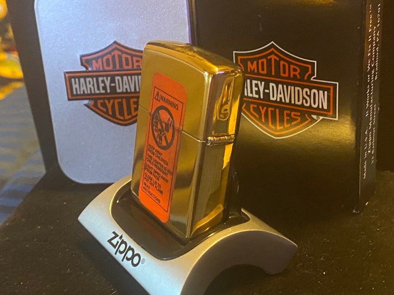 May include: A gold-tone Zippo lighter with an orange safety warning label on the front, resting on a silver Zippo display stand. Behind the lighter are two Harley-Davidson branded boxes, one silver and one black, featuring the iconic logo.