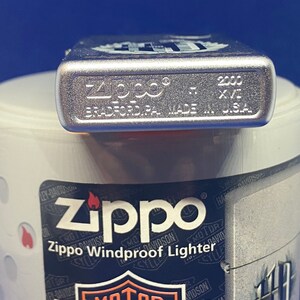 Puede incluir: Un encendedor Zippo Windproof plateado con el texto "Zippo" y "Bradford, PA. Made in U.S.A." impreso en la parte inferior. El encendedor est&aacute; sobre un recipiente blanco con un logotipo de Harley-Davidson.